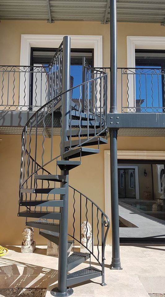 Escalier colimaçon gris dans une cour, marches ajourées et rampe à volutes